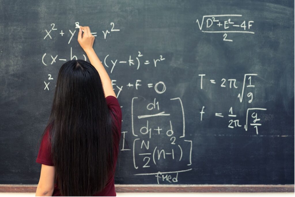Photo of a woman in from of a blackboard using chalk to write mathematical equations.