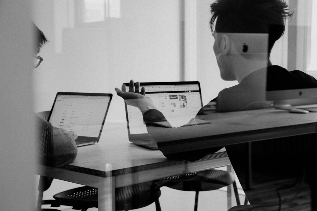 Two people discussing work while sitting at a desk using laptops.